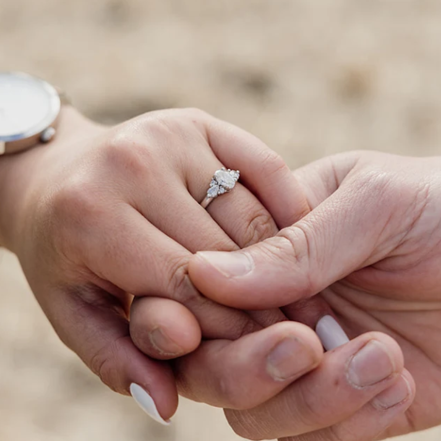 Close-up of two hands holding each other with a diamond ring on one finger, against a blurred natural background. Metal & Stone Happy Couple Testimony