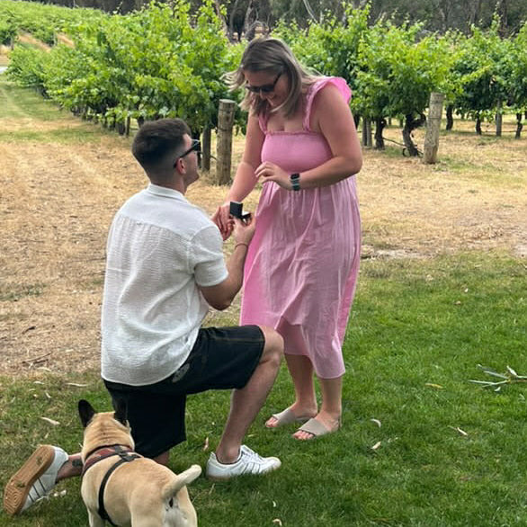 Man proposing to a woman in a pink dress in a vineyard with a dog present. Metal & Stone Happy Couple Testimony
